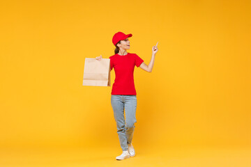 Full length body delivery employee woman in red cap blank t-shirt uniform work courier service in quarantine coronavirus covid-19 hold craft paper takeaway bag mockup isolated on yellow background.