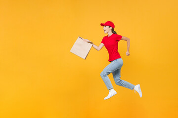 Full length jumping delivery employee woman in red cap blank t-shirt uniform work courier service in quarantine coronavirus covid-19 hold craft paper takeaway bag mockup isolated on yellow background