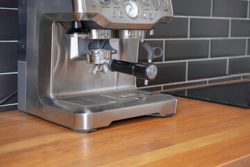 A carob type espresso machine stands on a wooden table top before grinding coffee into a filter holder against a black tile wall in a home kitchen. Making coffee at home.