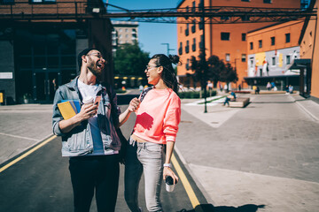 Cheerful male and female millennial students in stylish apparel emotionally laughing at urban setting during sunny summer day, prosperous diverse hipster guys enjoying live communication in city