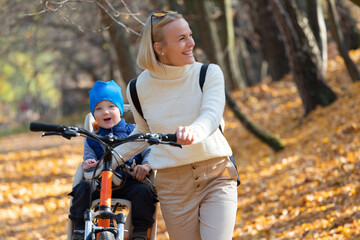 Happy mother leads a bicycle with a child strapped in the back in the autumn park.