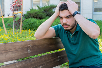 Young man with headache, holds his head with his hands. Young man with a beard sitting in a public park.