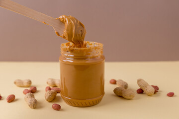 Peanut butter in a jar with nuts scattered on the table. Stretching peanut butter on a spoon. Colored background. Copy space for your text
