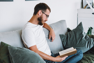 Fototapeta premium Young caucasian man resting on a sofa and reading a book. Home leisure.