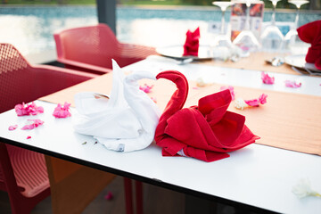 Red and white folded swan bird shape napkin in a restaurant on a white table at a resort near the pool. A flower petals and glasses on the table. Romantic dinner for newlyweds. Sunset.