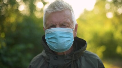 Portrait senior man puts on a white face mask to prevent the spread of the virus in city park or forest. Pandemic, Coronavirus. Senior man wearing anti virus mask is walking in the nature.