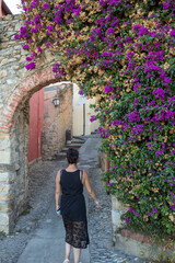 Woman with black dress strolls in an ancient village under a stone arch and flowers