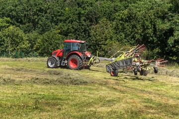 Obraz premium Red tractor working in a field with a two-rotor tedder. Drying hay in the meadow. Work on an agricultural farm.
