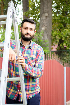 Young brunette bearded man standing on ladder on background of fence and trees in the backyard, wearing red green checkered shirt and looking at camera. Repair, yard work, gardening concept.Vertical
