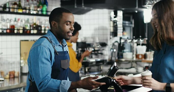 Multiethnic bartender man and client woman. Caucasian female paying with paypass, credit card. Mixed-races. Passing payment at counter. Pay for drink of coffee. African American waiter.