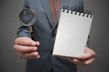 Magnifying glass and blank page notepad in the businessman hands close up on gray background.