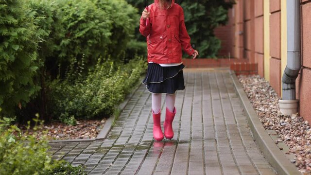 Happy kid girl in red rain coat and boots under umbrella enjoy jump and walk outdoors in first day of autumn