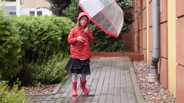 Happy kid girl in red rain coat and boots enjoy jump and spin around the umbrella walking outdoors in first day of autumn