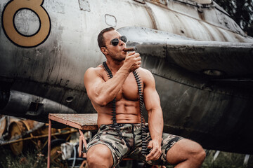Obraz na płótnie Canvas Old war airplane in the abandoned airport and brutal naked soldier near it. American bodybuilder smoking cuban cigar in the field.