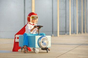 A super hero boy in a red helmet and a cape is driving a funny vehicle.