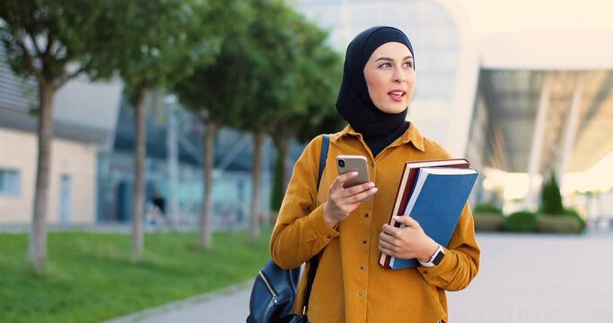 Young Arabic beautiful girl in hijab walking at street with textbooks, texting message on cellphone. Pretty muslim female student in headscarf strolling at city, holding books, tapping on phone.