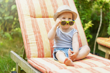 Smiling baby boy holding wearing sunglasses is sitting on the deck chair in the garden.