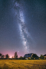 Fototapeta na wymiar Glowing milky way over crop field covered in fog. Starry night at farmstead. Farm house and tractor.
