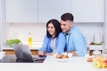 Obraz na płótnie Canvas Beautiful happy young couple having breakfast at home and uses laptop at kitchen. Enjoying free time together.