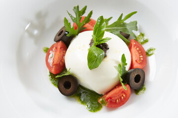 Traditional authentic soft and creamy fresh Italian burrata cheese shaped as white round ball over Italian tomatoes topped with rucola along side pesto reduction