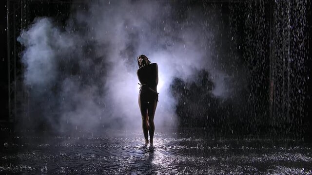Sensual erotic contemporary dance in the smoke among the raindrops. A young woman smoothly moves along the surface of the water, forming many splashes. Contemp choreography. Slow motion.