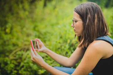 Side view of cute young brunette woman with glasses holding a white smartphone with red protective case in her hands and taking a picture. Green natural background and copy space on the left.