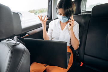 Quarantine time. Distant work. Business woman sitting in the car in the back seat wearing a protective mask and talks on the phone and working online using a laptop