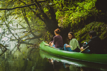 Group of three people, two young caucasian women and a man exploring, paddling with green canoe in...