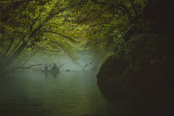 Fototapeta premium Back view of a green canoe and people in a misty and foggy river between the trees. Scary spooky and mistery exploration with a canoe on a river Krupa in Slovenia.