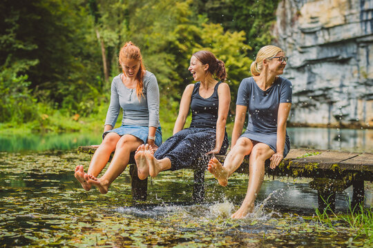 Three Young Women  Relaxing On A Wooden Pier And Splashing Their Feet Into Cold Water. Picturesque Clean Lake With Women Splashing Their Feet In Water. Concept Of Fun Sexy Outdoor Activity.