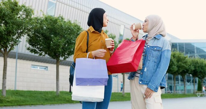 Two muslim stylish women in headscarves standing outdoor at city, talking and drinking coffee to-go. Stylish females at street chatting and sipping drink. Shopping. Shoppers. Best friends.