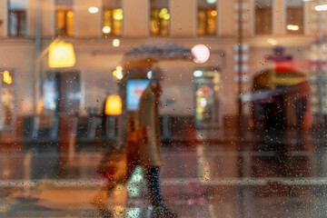 Blurred silhouette of an abstract unrecognizable girl with bags, view through a wet window with raindrops, city street in dark rainy evening