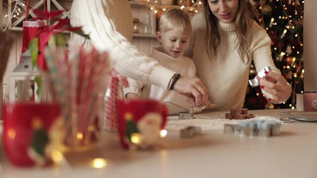 Happy beautiful family with a small blonde son roll out the dough for Christmas cookies in the kitchen, laughing. Dad smears his daughter's nose with flour. Slow motion. High quality HD footage