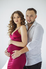 A beautiful curly pregnant girl in an evening dress stands in an embrace with her husband on a white background. She has a crimson evening dress and he is wearing festive white shirt. Family Concept