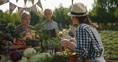 Senior caucasian couple selling fresh local fruits and vegetables from their ranch. Young female customer buying vegetarian groceries at farmers market 4k footage - Powered by Adobe