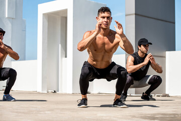 Group of fit sports men doing squat bodyweight workout training outdoors on building rooftop -...
