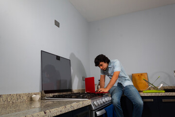 Young hispanic wavy hair man studying from home with a laptop in the kitchen whether sitting or...