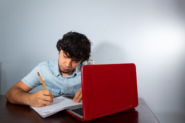 Young hispanic wavy hair man studying from home with a laptop and writing with a pencil in a notebook