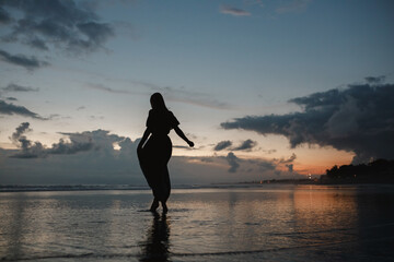 Silhouette woman in long dress walks on the sandy beach and enjoying the sunset, twilight