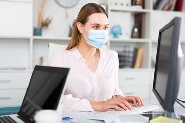 Focused young woman in disposable face mask working in business office using laptop
