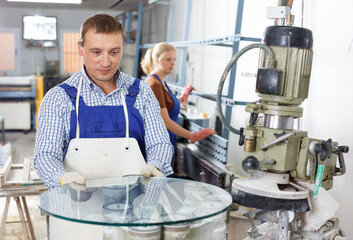 Serious craftsman working on machine for processing of curved facet of glass in workshop.