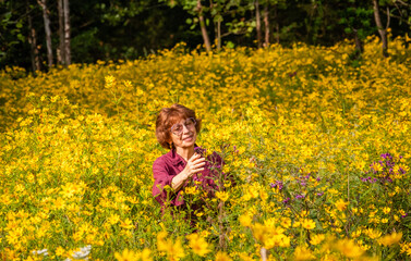 Senior woman enjoying tall yellow flowers growing in field  in early autumn; woods in background