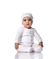 Cute infant baby toddler sits on the floor in white jumpsuit overall and funny hat with ears holding hands at knees