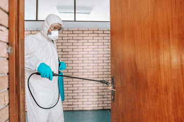 Worker in white sterile uniform, with rubber gloves and mask on holding sprayer with disinfectant and sterilizing doors in school.