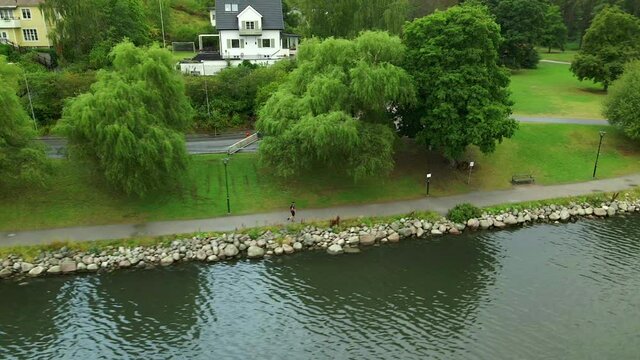 A female running a long sida of a small lake with trees and a white house in the background. Orbiting aerial view.