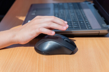 A child's hand holds a computer mouse next to a laptop keyboard on a wooden table.
