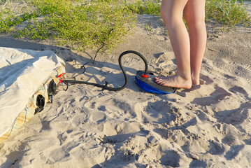 A woman with air foot pump pumps an inflatable mattress or air bed at sandy beach. Foot inflates air mattress with foot pump on sand.