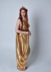 Full length portrait of girl with red hair wearing long grecian toga and golden wreath. Standing pose iisolated against a grey studio background. - obrazy, fototapety, plakaty
