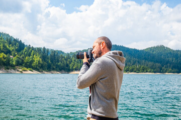 Photographer man in hoodie walking around lake with DSLR camera and shooting nature, half body portrait, landscape photography concept
