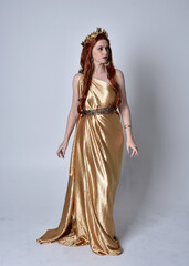 Fototapeta premium Full length portrait of girl with red hair wearing long grecian toga and golden wreath. Standing pose iisolated against a grey studio background.